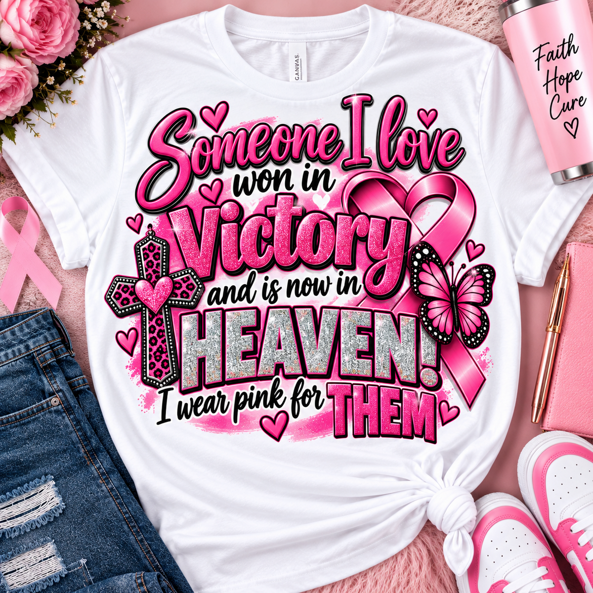 White graphic T-shirt laid flat with a pink and glitter memorial-style message reading 'Someone I love won in Victory and is now in HEAVEN! I wear pink for THEM,' surrounded by pink hearts, a ribbon, cross, butterfly, flowers, denim shorts, a pink Hope Care tumbler, a notebook, and pink sneakers on a pink textured background.